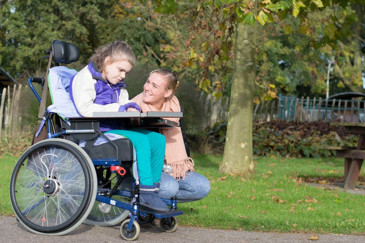 Girl in wheelchair with caring woman outside.