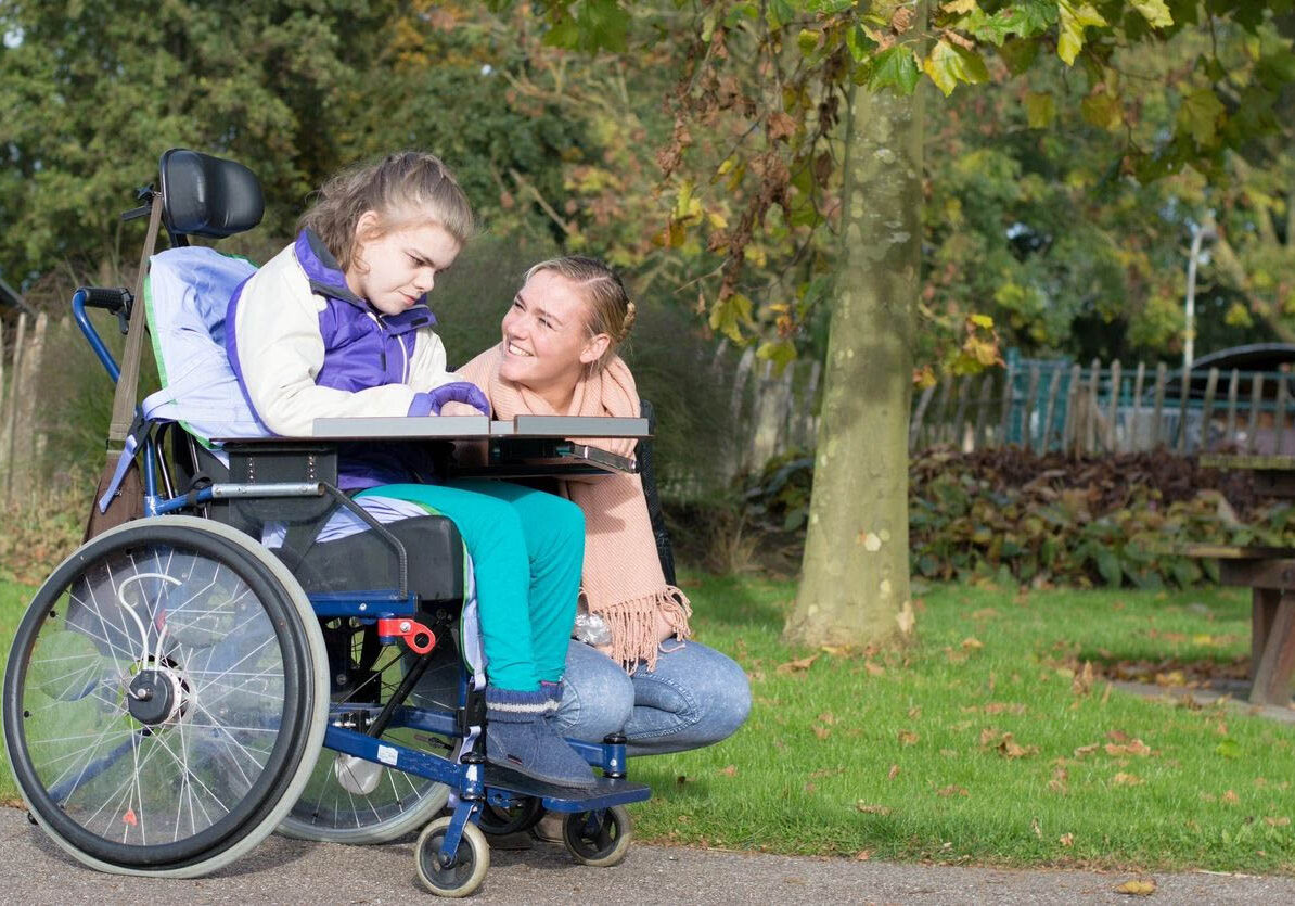Girl in wheelchair with caring woman outside.